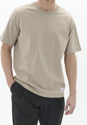 Beige T-shirt med korte ærmer, rund halsudskæring, glat tekstur og en lille hvid label ved kanten. Kombineret med mørke bukser.