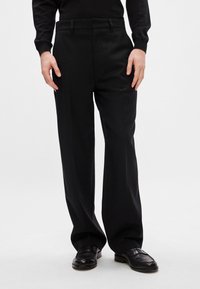 Calças pretas ajustadas com corte reto, apresentando uma textura suave e bolsos laterais. Combinadas com mocassins de couro pretos.