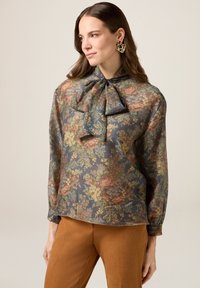 Blommig blus i mörkblått, med en rosett vid kragen, långa ärmar och ett genomskinligt, strukturerat tyg med färgglada blommönster.