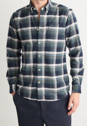 Man wearing navy, green, and white plaid button-up shirt with long sleeves and navy pants, standing with one hand in pocket.