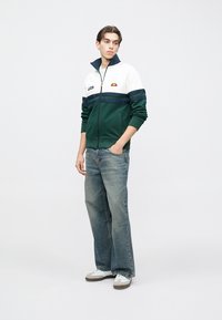 Zaļa un balta rāvējslēdzēja jaka ar augstu apkakli, ar tumši ziliem horizontāliem svītrām un nelielu logotipu. Valīta ar gaišām džinsa biksēm un sporta apaviem.