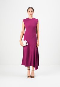 Donna che indossa un vestito midi viola asimmetrico senza maniche, tacchi neri con fascette, mentre tiene una piccola borsa bianca arricciata, in piedi davanti a uno sfondo bianco.