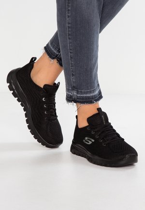 Stopy noszące czarne dziane tenisówki Skechers z czarnymi podeszwami i sznurowadłami, w połączeniu z ciemnoszarymi dżinsami ze strzępionym dołem na białej podłodze.