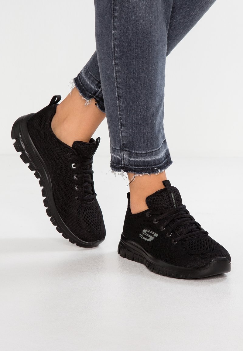 Pieds chaussés de baskets tricotées noires Skechers avec semelles et lacets noirs, associées à un jean gris foncé à ourlet effiloché sur un sol blanc.