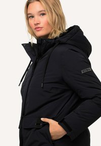 Chaqueta aislante negra con capucha, con cremallera frontal, cordones ajustables y un parche de la marca en el brazo izquierdo.
