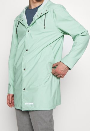 Personne portant un imperméable à capuche vert clair avec boutons-pression et la main dans la poche latérale, associé à un pantalon gris.