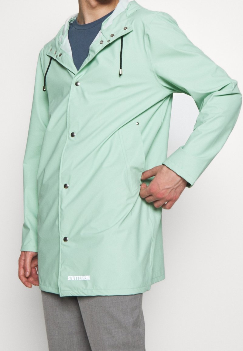 Personne portant un imperméable à capuche vert clair avec boutons-pression et la main dans la poche latérale, associé à un pantalon gris.