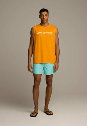 Pomarańczowa bezrękawnik z napisem "różnorodność", w połączeniu z jasnoniebieskimi szortami i czarnymi japonkami. Prosta, casualowa letnia stylizacja.