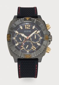 Svart gummireim klokke med mørkegrå klokkehus, gullakenter, teksturert svart urskive og tre subskiver. Har en tachymeter bezel.