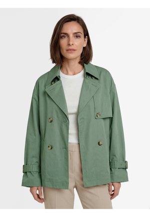 Frau mit kurzem braunem Haar, die eine hellgrüne, doppelt geknöpfte Jacke über einem weißen Hemd und beigen Hosen trägt, steht vor einem schlichten Hintergrund.