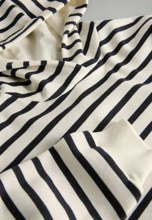 Valge ja musta horisontaalse triibuga kangas lähivõte, kus on näha kapuuts ja soonikkoes mansett pikkade varrukatega rõival.