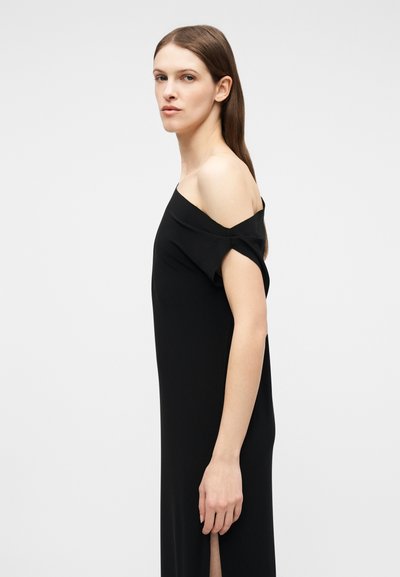 Γυναίκα με μακριά καστανά μαλλιά, ντυμένη με μαύρο φόρεμα χωρίς ώμους, που στέκεται πλάγια μπροστά σε απλό φωτεινό φόντο.