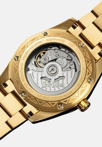 Goldfarbene Uhr mit einem strukturierten Gehäuseboden, sichtbarem mechanischen Uhrwerk, Saphirglas und graviertem Markennamen.