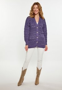 Cardigan tricotat violet cu decolteu în V, model texturat și nasturi mari, purtat peste un top alb, asortat cu pantaloni deschiși și cizme bej.