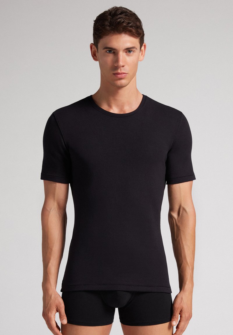 Jonge man die frontaal staat, draagt een aansluitend zwart T-shirt met korte mouwen en zwarte boxershorts tegen een effen achtergrond.