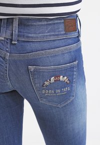 Blå denimjeans med en blekt finish, designade med en detaljerad bakfickedesign med broderad text och loggopunkter.
