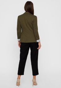 Olivgrön skräddarsydd blazer med en figurnära passform, kombinerad med svarta byxor. Har en subtil textur och manschettdetaljer vid ärmarna.