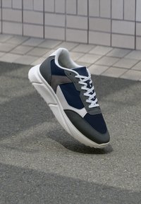 Dunkelblaue und graue Sneakers mit weißer Sohle und Schnürsenkeln, schräg auf Asphalt neben einer gefliesten Wand positioniert.