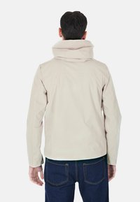 Giacca impermeabile beige con un collo alto e cappuccio, caratterizzata da una texture liscia e da una silhouette aderente, abbinata a jeans blu scuro.