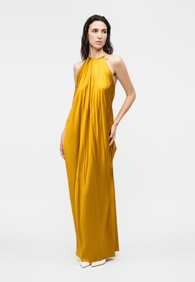 Γυναίκα με μακριά σκούρα μαλλιά που φορά ένα κίτρινο μουσταρδί φόρεμα μέχρι το πάτωμα χωρίς μανίκια και άσπρα μυτερά παπούτσια, στέκεται με το ένα χέρι στη μέση.