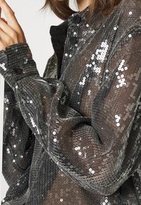 Giacca nera con paillettes e un layer esterno trasparente, dotata di polsini con bottoni, colletto e una superficie texturizzata che riflette la luce.