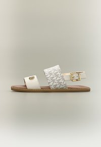 Λευκό φλατ σανδάλι με δύο λουριά, το ένα με χρυσό έμβλημα και το άλλο με σχεδιασμένη μοτίβα, συν λουρί αστραγάλου με χρυσή αγκράφα.