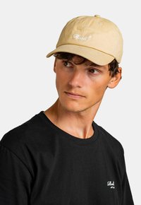 Beige Baseballkappe mit gesticktem weißen Text, getragen mit einem schwarzen T-Shirt. Die Kappe hat einen gebogenen Schirm und einen verstellbaren Riemen auf der Rückseite.