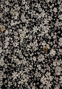 Un tessuto nero presenta un motivo floreale bianco con diverse forme di fiori. Due bottoni dorati sono visibili nella zona della tasca sul petto.