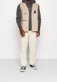 Gilet utility beige con multiple tasche, indossato sopra un felpa grigia. Pantaloni chiari e sneakers nere completano l'outfit.