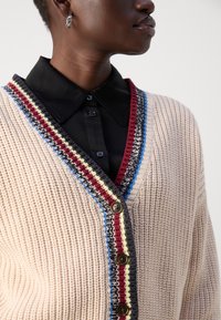 Cardigan beige lavorato a maglia con scollo a V profondo, caratterizzato da un bordo a strisce multicolori e bottoni marroni. Indossato sopra una maglietta nera.