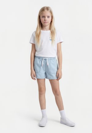 Blond dievča stojí, má na sebe biele tričko, svetlomodré šortky s šnúrkou, biele ponožky a biele slip-on topánky na jednomfarbenom pozadí.