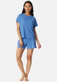 Frau steht barfuß, trägt ein blaues kurzärmeliges Oberteil und passende Shorts, lächelt mit entspannter Haltung vor weißem Hintergrund.