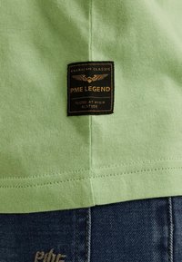 T-shirt en coton vert clair avec un ourlet cousu, arborant une étiquette rectangulaire noire inscrite "PME LEGEND" et un design d'aigle.