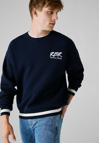 Granatowy sweter z białym haftem "RBR" i "RED BULL RACING". Posiada prążkowany okrągły dekolt oraz paskowane mankiety. Noszony z jasnoniebieskimi dżinsami.