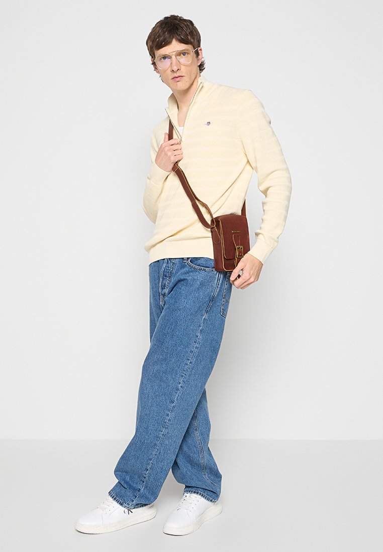 Jeune homme portant un pull beige à col zippé, un jean bleu décontracté, des baskets blanches, des lunettes transparents et un sac bandoulière marron, se tenant devant un fond blanc.