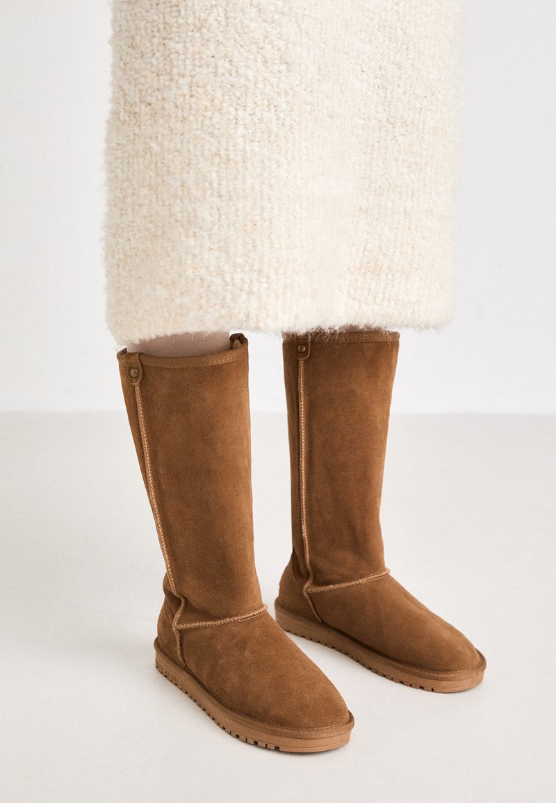 Bottes en suède marron à hauteur de genou avec une texture lisse, présentant des coutures latérales et une semelle en caoutchouc pour une bonne traction.