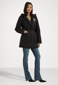 Quiltad svart jacka med huva, framfickor och en gyllene logotyp. Den har en figursydd design med midjeband och ribbade ärmslut.