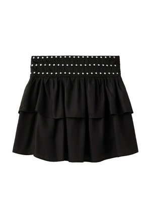 Femme portant une robe noire sans manches à volants avec une taille cloutée et des sandales plates noires à brides cloutées, debout sur fond blanc.