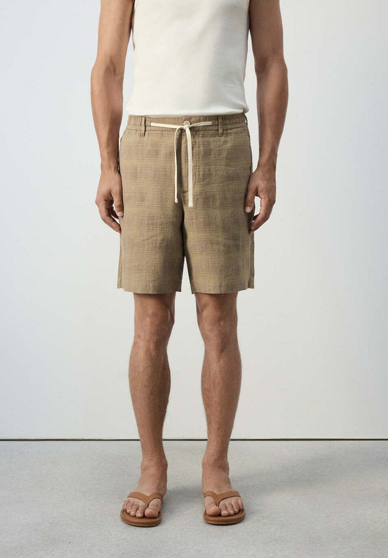 Homme portant un short beige à cordon, un débardeur blanc et des sandales marron, debout devant un fond clair uni.