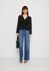 Schwarze Bluse mit Wickeldesign und Spitzenakzenten an den Schultern, kombiniert mit hellblauen weit geschnittenen Jeans, schwarzer Tasche und Sandalen mit Riemchen.