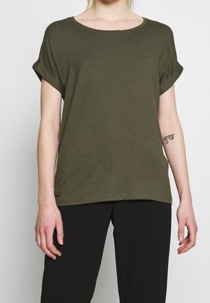 Person iført en olivengrøn kortærmet T-shirt med opkrogede ærmekanter og sorte bukser, der viser en lille tatovering på venstre underarm.