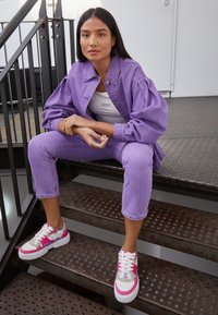 Jaqueta de ganga lavanda com mangas bufantes, calças cropped lavanda a condizer e ténis com pormenores em rosa. Fundo industrial com escadas de metal.