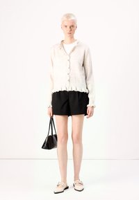Cardigan crema con bottoni e orlo arricciato, pantaloni corti neri, borsa nera, scarpe beige con cinturini, in piedi su uno sfondo bianco.