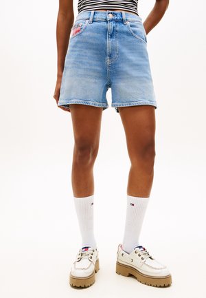 Άτομο που φοράει ανοιχτό μπλε τζιν σορτς, λευκές κάλτσες μέχρι τη μέση της γάμπας και χοντρά λευκά παπούτσια με ταμπά σόλες, στέκεται μπροστά σε μια απλή φόντο.