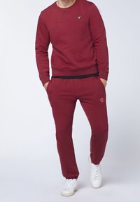 Sudadera burdeos con cuello redondo y puños acanalados, combinada con pantalones de chándal burdeos a juego. Presenta un logotipo y una cinturilla elástica. Completado con zapatillas blancas.