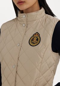 Béžová prošívaná vesta s vysokým límcem, se stříbrnými druky a vyšívaným emblémem se zlatými akcenty.