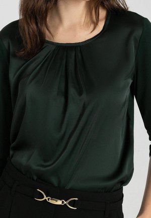 Frau trägt eine dunkelgrüne seidige Bluse, die in schwarze Hosen gesteckt ist, mit einem schwarzen Gürtel mit goldener Metallschnalle.