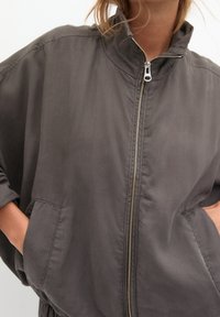 Vrouw in een grijze zip-up jas met hoge kraag en handen in de zijzakken, cropped onder de neus en boven de heupen.