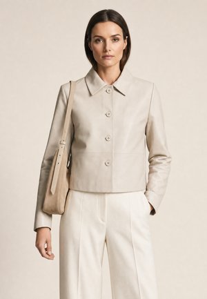 Frau trägt eine beige Jacke mit Knöpfen, hoch taillierte cremefarbene Hose und eine Schultertasche, steht vor schlichtem Hintergrund.
