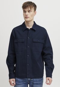 Veste boutonnée bleu marine en coton avec deux poches poitrine, manches longues et col. Tissu texturé au design classique.
